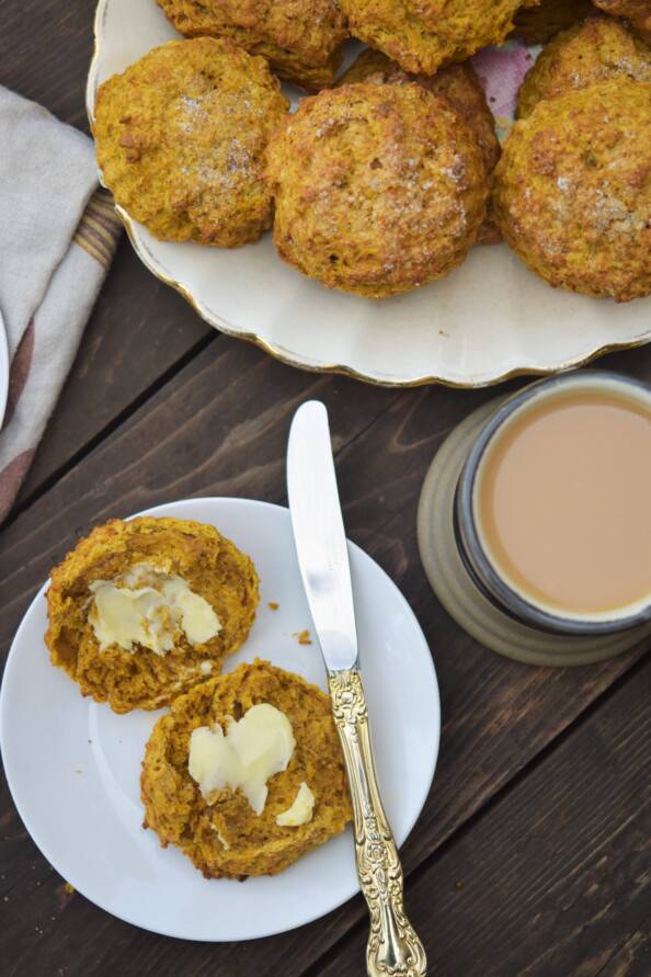 overhead shot of a buttered pumpkin scone on a white plate with a cup of tea and platter of scones