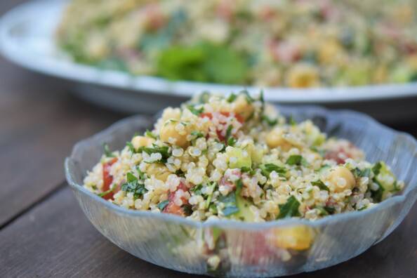 A serving of quinoa tabbouleh salad in a clear glass bowl, with a larger platter of it in the background